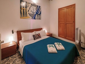 A hotel room with a bed, a wooden headboard, a nightstand with a lamp, a wall-mounted artwork, a wooden wardrobe, and a folded towel on the bed.