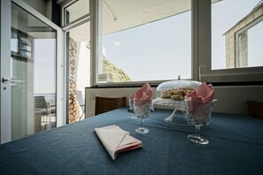 A table with a blue tablecloth, two wine glasses, and a glass bowl with food items, set against a backdrop of a window with a view of a hillside.