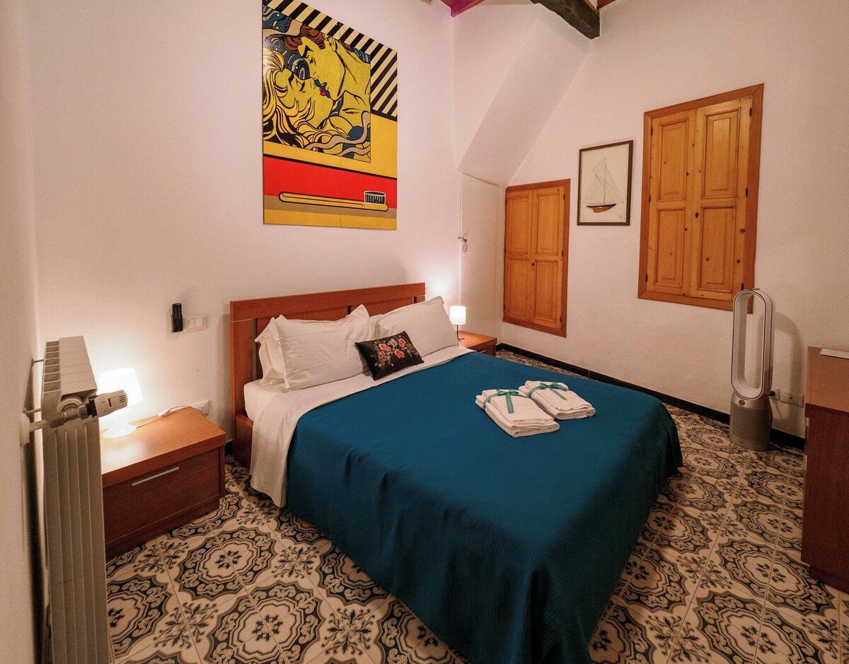 A hotel room with a bed, a nightstand, a radiator, a wall-mounted artwork, a wooden door, and a window with a sailboat.