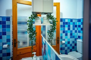 Une salle de bain avec des carreaux bleus, des toilettes, un lavabo avec un distributeur de savon et une fenêtre avec des stores.