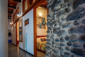 Un couloir avec un mur en pierre, des poutres en bois, un radiateur et un lit recouvert d’une couvre-lit à motifs floraux.