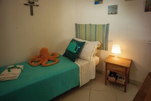 A single bed with a teal bedspread, an orange plush octopus, a pillow, and a bedside lamp on a wooden nightstand.