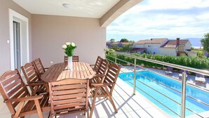 En balkon med et træbord og stole, en blomstervase og udsigt til en swimmingpool og huse i baggrunden.