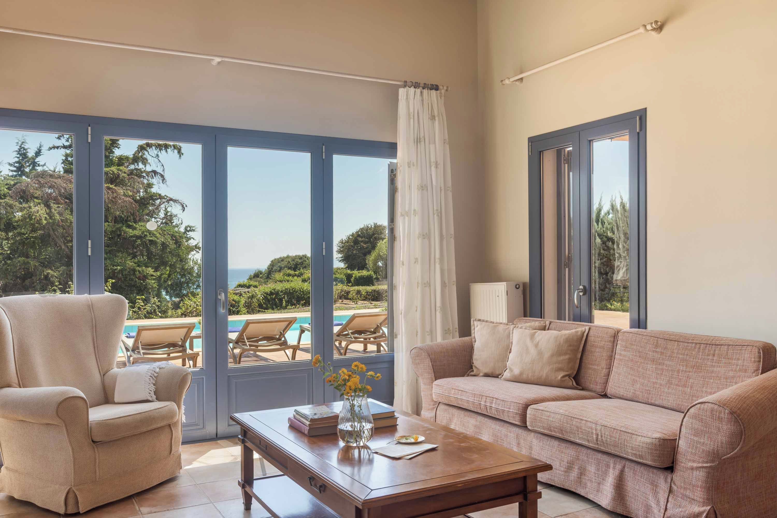 A living room with a sofa, armchair, and a coffee table. There is a view of a pool and greenery outside.