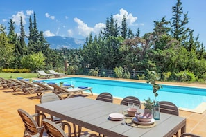 A poolside area with a dining table set for four, surrounded by lounge chairs, and a view of a mountain range in the background.
