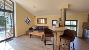 Un salon moderne avec un canapé, une table basse et deux tabourets de bar. Une grande fenêtre laisse entrer la lumière naturelle et offre une vue sur l’extérieur.