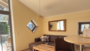 Un salon moderne avec un canapé, une table ronde et une chaise. Il y a une fenêtre donnant sur des arbres et un balcon.