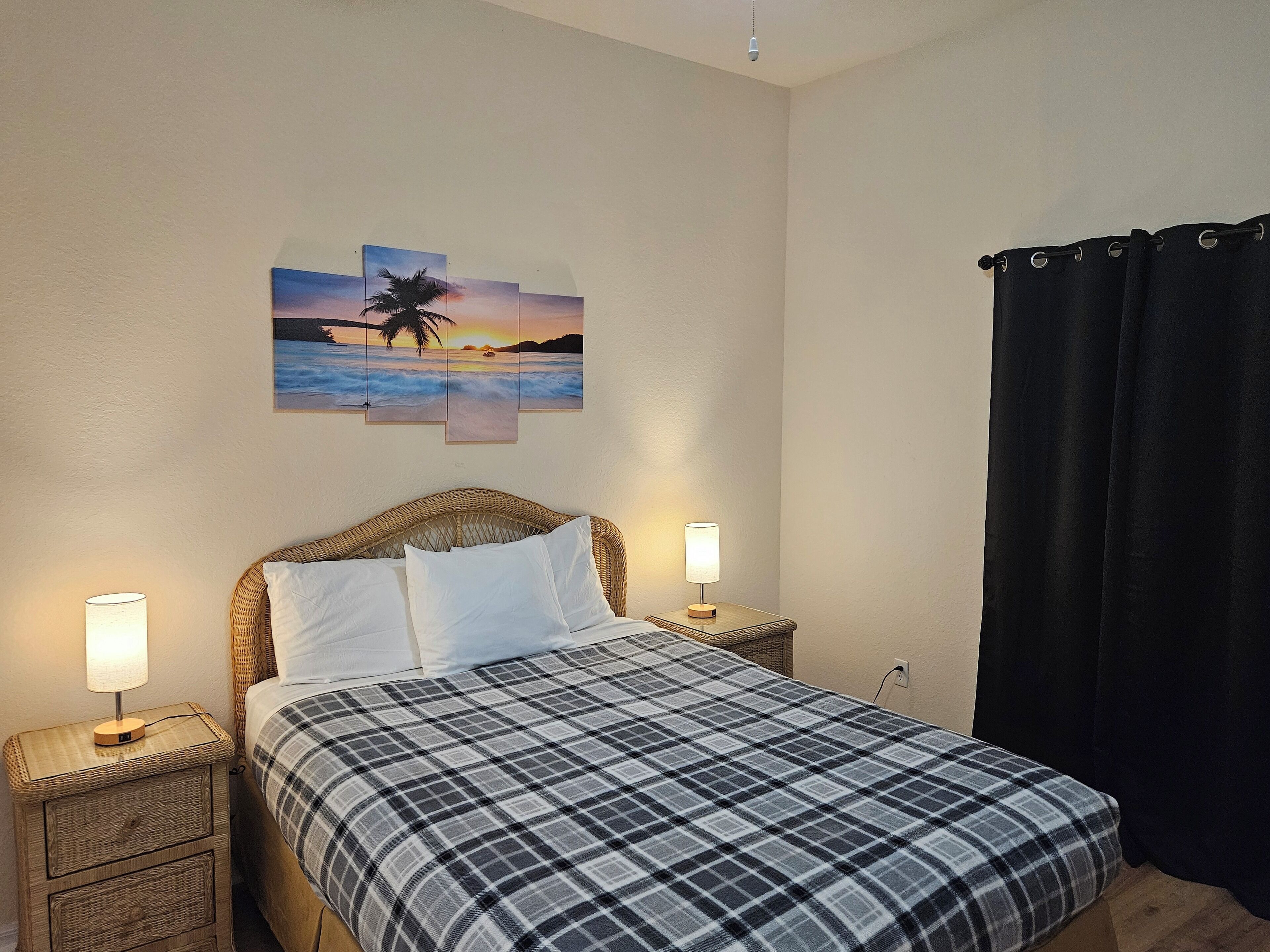A bedroom with a bed, two bedside tables with lamps, a wall-mounted painting, and a black curtain.