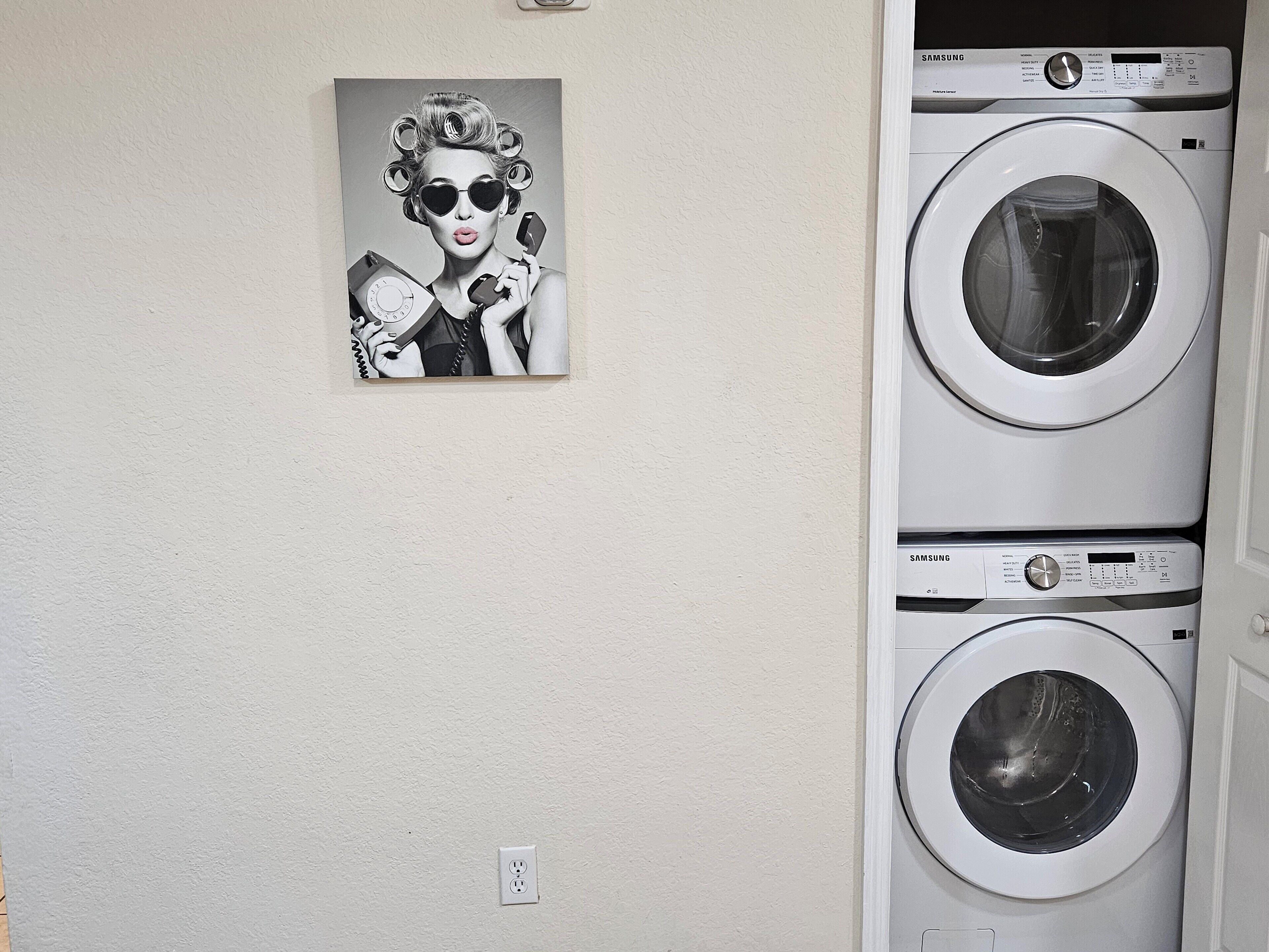 A wall-mounted Samsung washing machine with a control panel, a power outlet, and a black and white photograph of a woman with curlers and a rotary dial telephone.