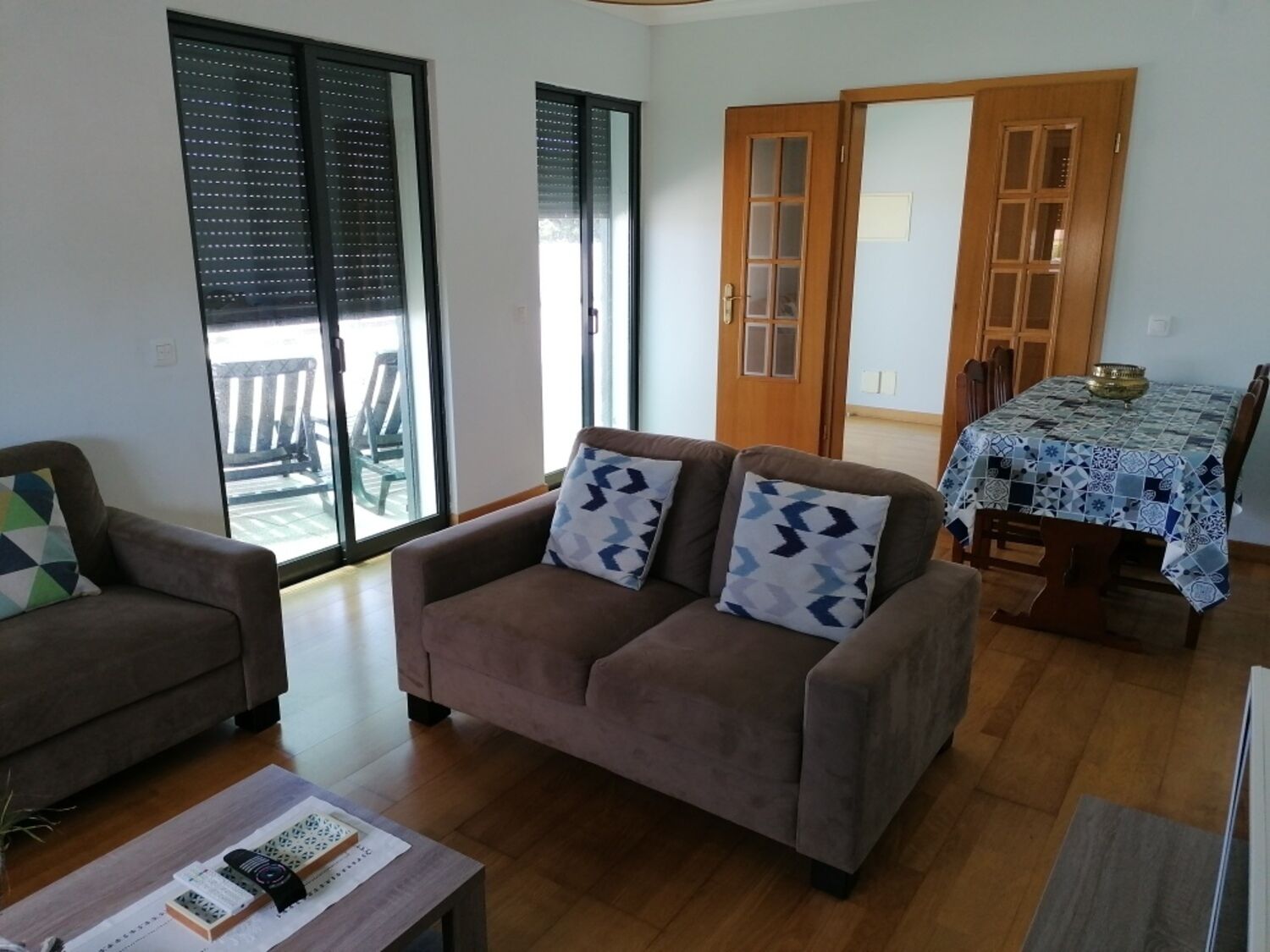 A living room with a sofa, a coffee table, and a dining table with chairs. There is a sliding glass door leading to an outdoor area with chairs.