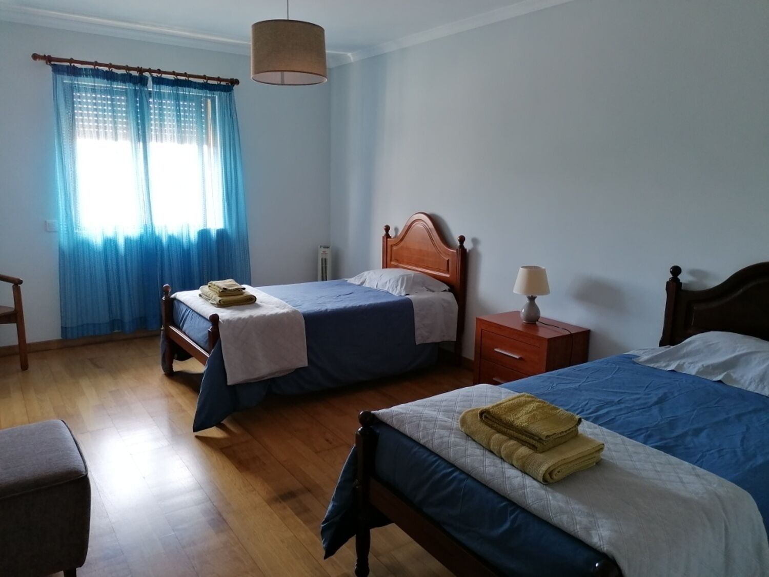 A bedroom with two beds, a wooden floor, a window with blue curtains, a bedside table with a lamp, and a chair.