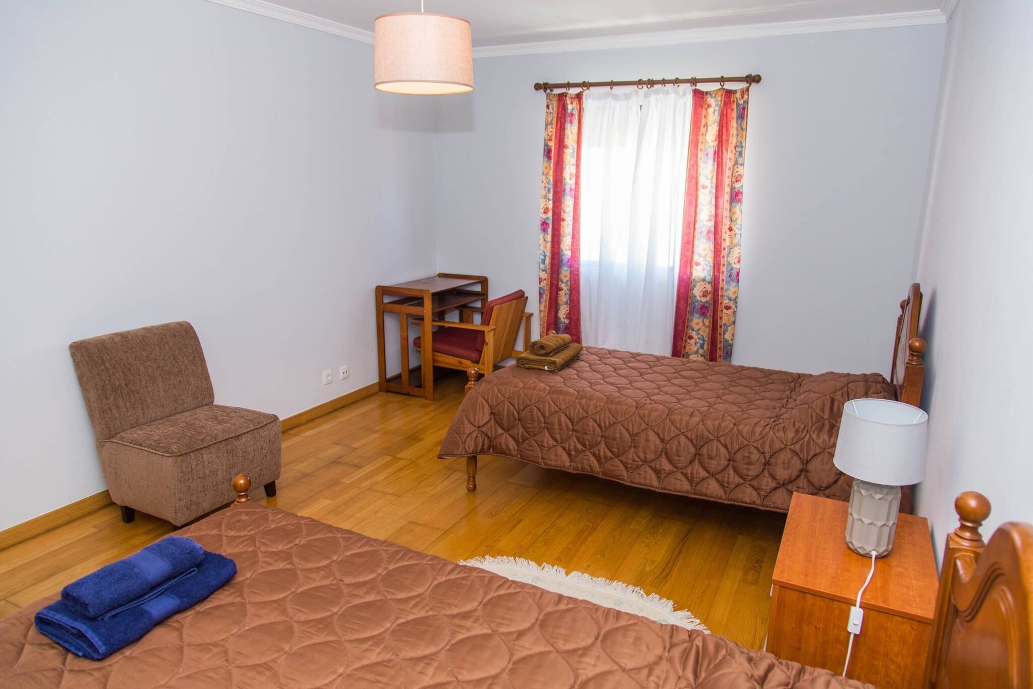 A bedroom with two beds, a chair, a small table, a lamp, and a window with curtains.