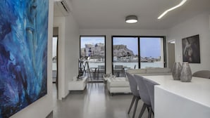 Un salon moderne agrémenté d’un grand tableau abstrait, un coin repas avec une table et des chaises blanches, et de grandes fenêtres offrant une vue sur un paysage urbain et une étendue d’eau.