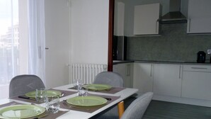Ein Essbereich mit einem Tisch, gedeckt für vier Personen, einschließlich Tellern, Besteck und Gläsern. Stühle umgeben den Tisch, und im Hintergrund ist eine Küche erkennbar.