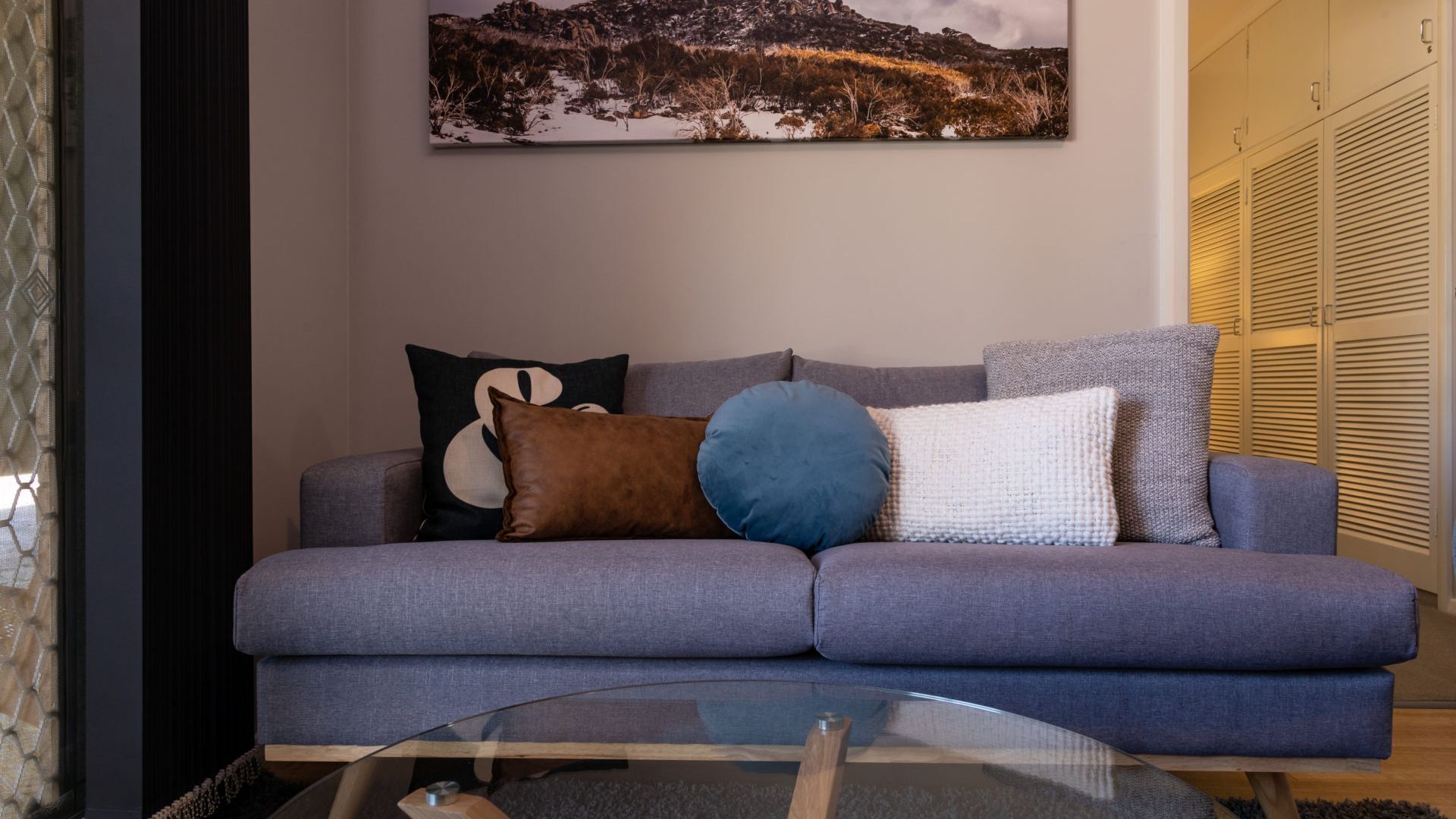 Una sala de estar con un sofá gris, una mesa de centro de vidrio y un cuadro de un paisaje en la pared.