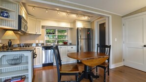 A kitchen with white cabinetry, a stainless steel refrigerator, a dining table with chairs, and a window with a view of greenery. - Blue Zen overlooking nature filled buoy beauty! (Saugatuck)