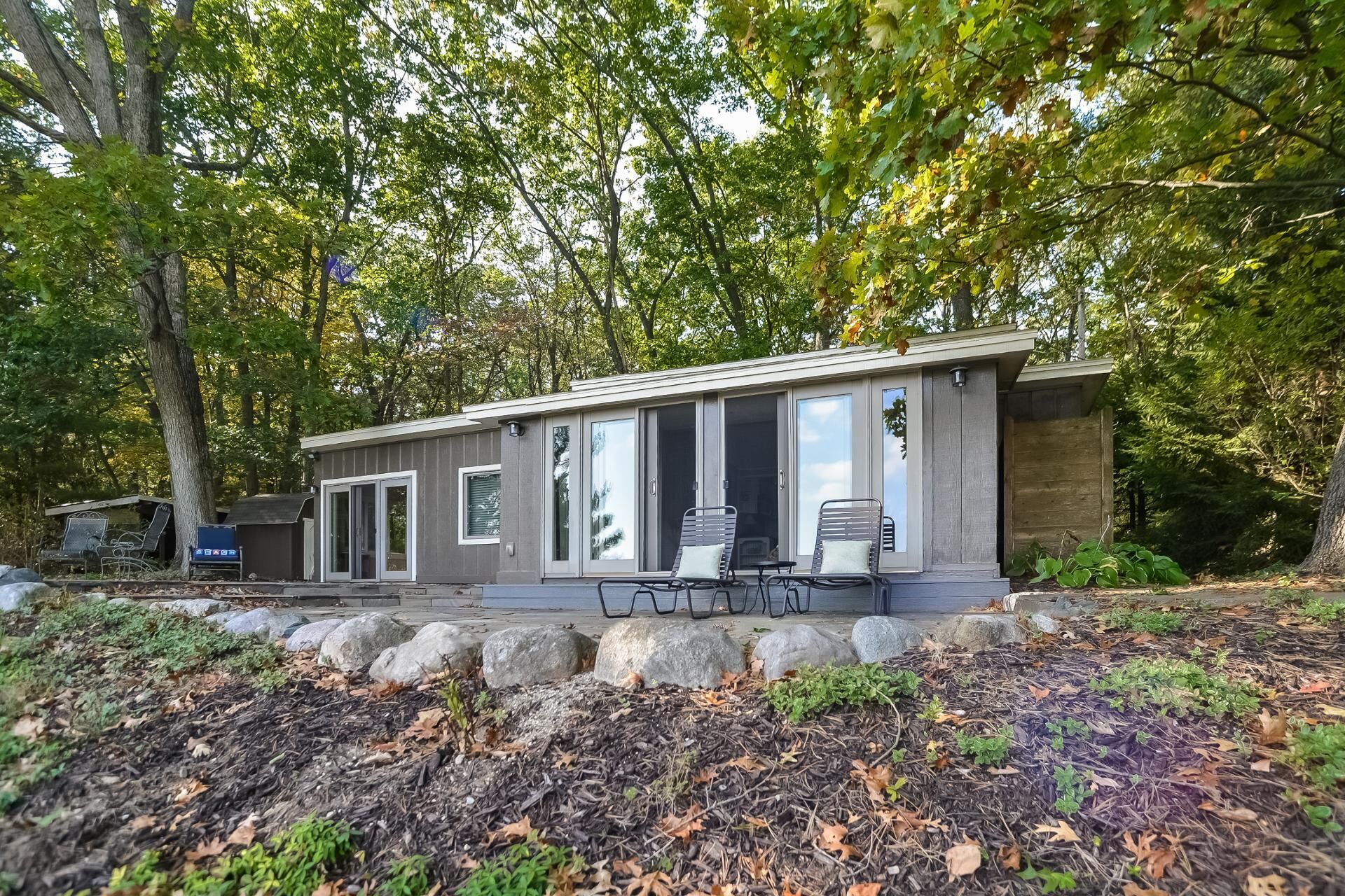 A modern cabin with large sliding glass doors, a patio with outdoor seating, and a stone retaining wall.