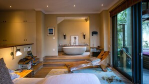 A spacious bedroom with a large bed, a wooden bench, and a view of a large bathtub in the background. - TABLE MOUNTAIN VILLA (Newlands)