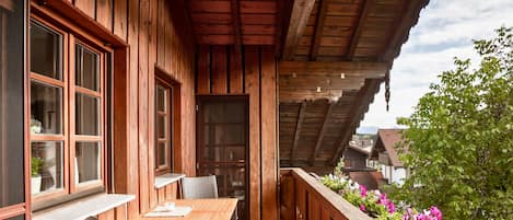 A wooden balcony with a table and chairs, overlooking a garden with pink flowers.