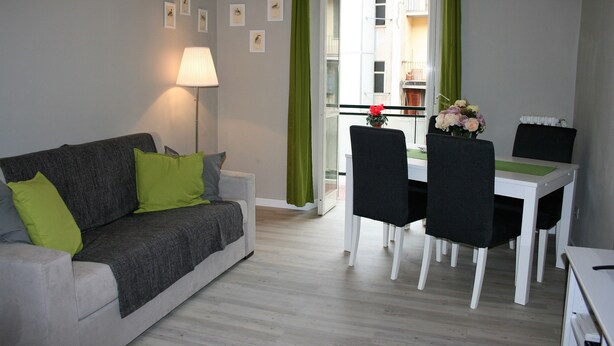 Un salon avec un canapé gris, des coussins verts, une table basse blanche et un coin repas comprenant une table blanche et des chaises noires.