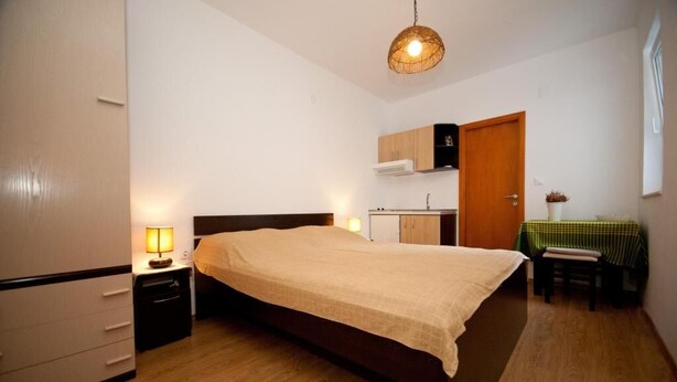 A bedroom with a bed, bedside table, lamp, and a small table with a green tablecloth. - Apartments Briest - Studio Apartment (Dubrovnik)