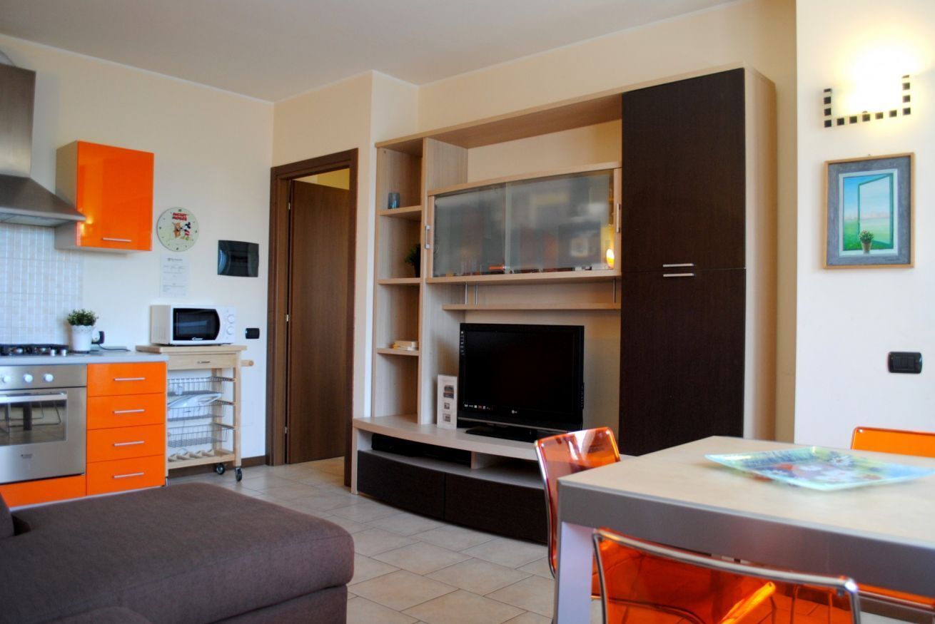 Une cuisine moderne équipée d’un four à micro-ondes, d’une cuisinière et de meubles de rangement orange. On y trouve également un coin repas avec une table et des chaises, ainsi qu’un téléviseur posé sur une étagère en bois.