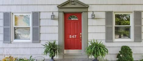 Una casa de dos pisos con una puerta roja, número 547, y un pequeño porche con plantas en macetas.