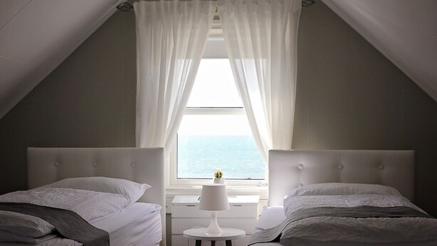 A bedroom with two beds, a small table, and a window with curtains. - A House Between Mountains and Coast in a Beautiful and Quiet sea Village (Skagaströnd)