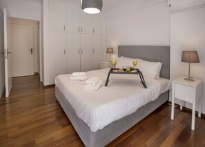 A neatly made bed with white linens and a tray of breakfast items, including fruits and juice, in a modern hotel room with wooden flooring and white furniture.