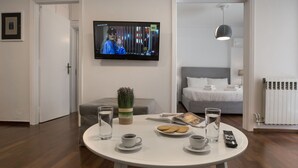 A modern living room with a round white table, a flat-screen TV mounted on the wall, a grey sofa, a grey bed, and a white radiator.