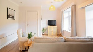 Un salon moderne comprenant un canapé, un fauteuil, une petite table avec une plante, un téléviseur posé sur un meuble en bois et une grande fenêtre avec des rideaux.