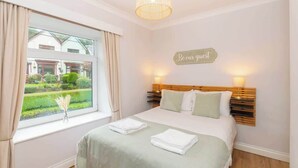 Une chambre d’hôtel avec un lit, une fenêtre avec des rideaux, un panneau sur le mur et une lampe sur la table de chevet.