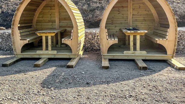 Deux structures en bois en forme d’arche, dotées de bancs, qui semblent être des espaces de repos aménagés à l’extérieur.