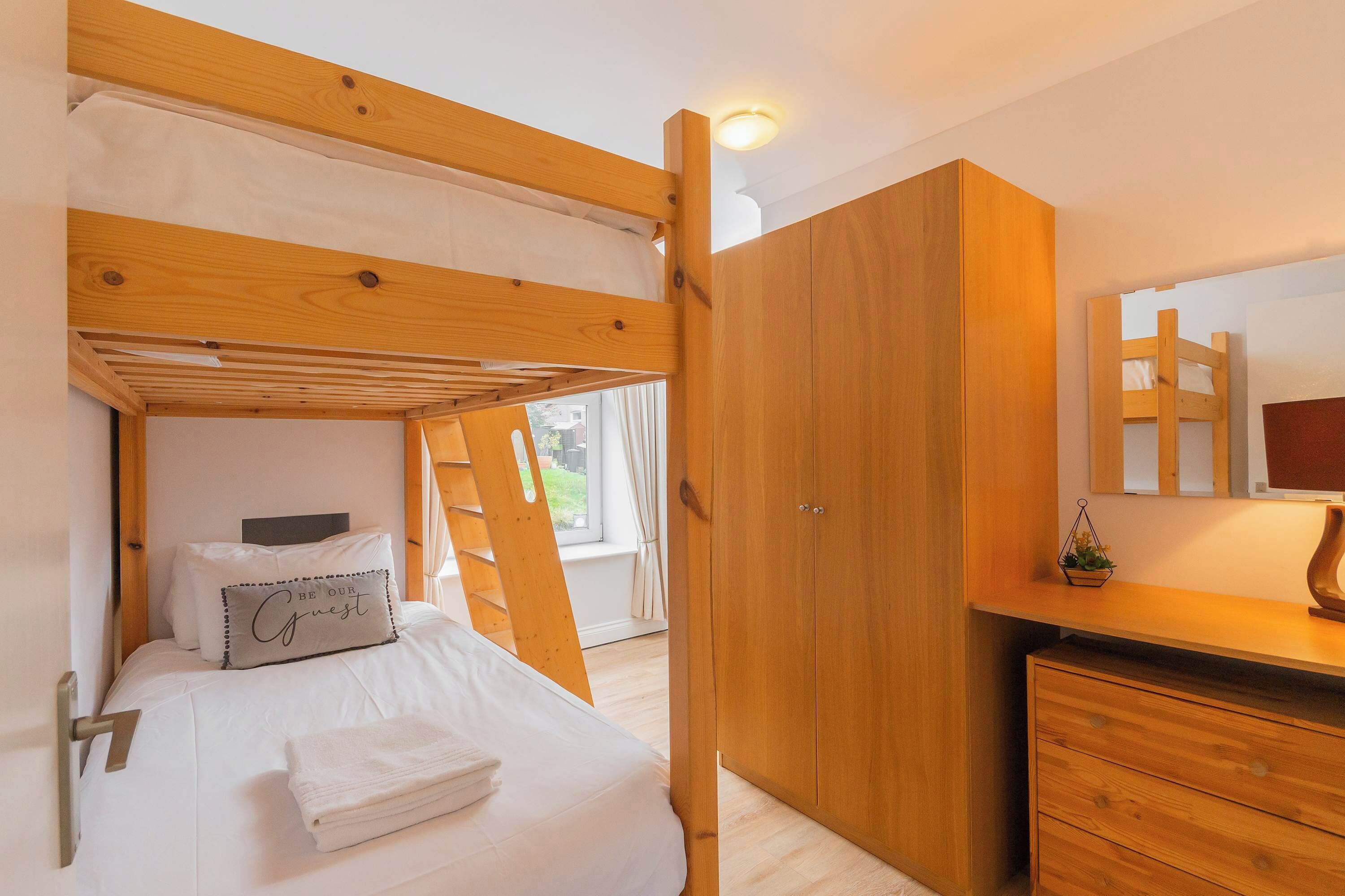 A bunk bed with a wooden headboard, a bedside table with a lamp, a wardrobe, and a small plant on the dresser.