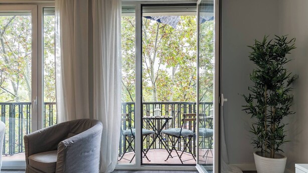 Ein Zimmer mit einer Schiebetür zu einer Terrasse im Freien, mit Tisch und Stühlen. Im Zimmer steht ein Blumentopf und ein gepolsterter Sessel.