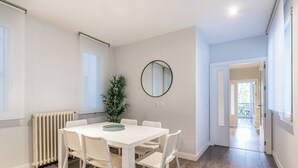 Ein zeitgemäßer Essbereich mit einem weißen Tisch und Stühlen, einer Topfpflanze und einem runden Spiegel an der Wand.