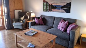 A cozy living room with a grey sofa, a wooden coffee table, a grey armchair, and a lamp. A painting of a mountain landscape hangs on the wall. - Vacation apartment Birnbaum 68 sqm, 2 separate bedrooms, kitchen (Neubeuern)