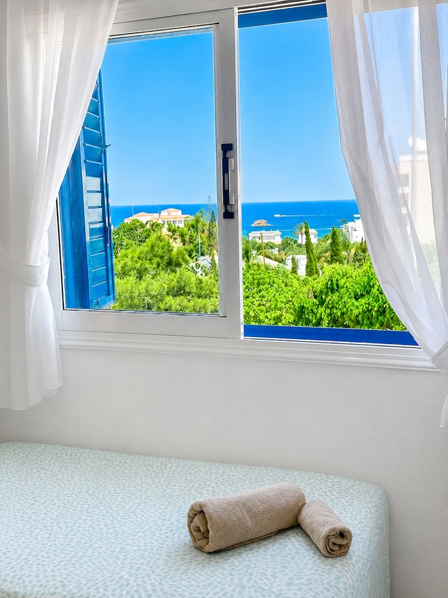A room with a bed, a window with white curtains, and a view of the sea and greenery.