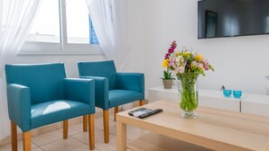 Uma sala de estar moderna com duas poltronas azul-turquesa, uma mesa de centro de madeira, um vaso com flores, uma TV de tela plana e uma janela com cortinas leves.