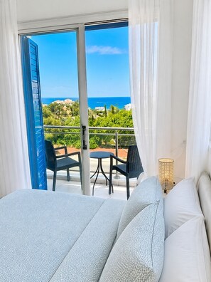 Une chambre d’hôtel avec balcon, un lit avec une literie blanche et une vue sur la mer et la verdure.