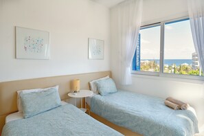 A hotel room with two beds, a small table, and a window with a view of the sea and buildings.