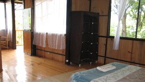 A room with two beds, wooden floors, a wardrobe, and a view of greenery outside.