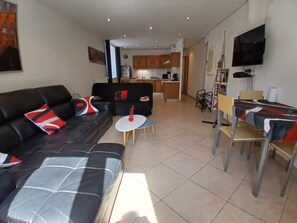 A modern living room with a sofa, a dining table with chairs, a kitchen in the background, and a TV mounted on the wall.