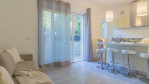 Een moderne woonkamer met een bank, een keukenhoek met barstoelen en een schuifpui naar een balkon.
