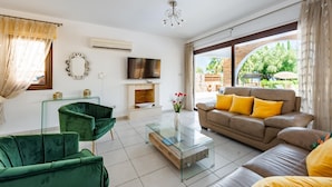A modern living room with a green sofa, a beige sofa with yellow cushions, a glass coffee table, a flat-screen TV, and a fireplace.
