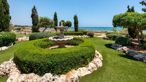A well-manicured garden with a central fountain, neatly trimmed hedges, and a view of the sea.