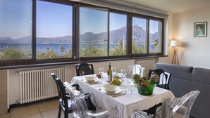 Un coin repas avec une table dressée pour quatre personnes, des chaises transparentes, une vue sur la mer et les montagnes, un radiateur et une lampe.