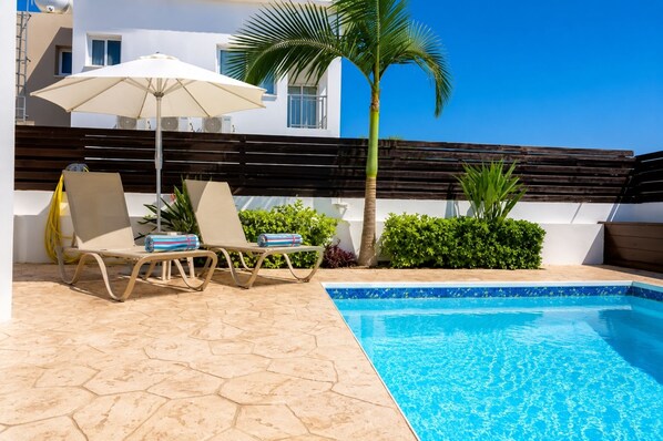 A swimming pool with lounge chairs and an umbrella, surrounded by greenery and a wooden fence.