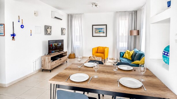 A modern living room with a dining area, featuring a wooden dining table set for four, a sofa with yellow cushions, and a television on a wooden cabinet. - Narcissos Villa Doria (Protaras)