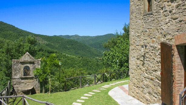 Ein Steinhaus mit einem kleinen Fenster und einer Holztür, umgeben von einer Steinmauer und einem Metallgeländer.